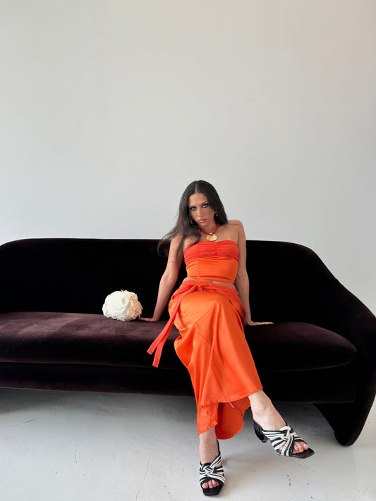 Model wearing a satin strapless bustier top and matching satin maxi skirt with a chiffon waist tie, posing against a minimalist studio backdrop.