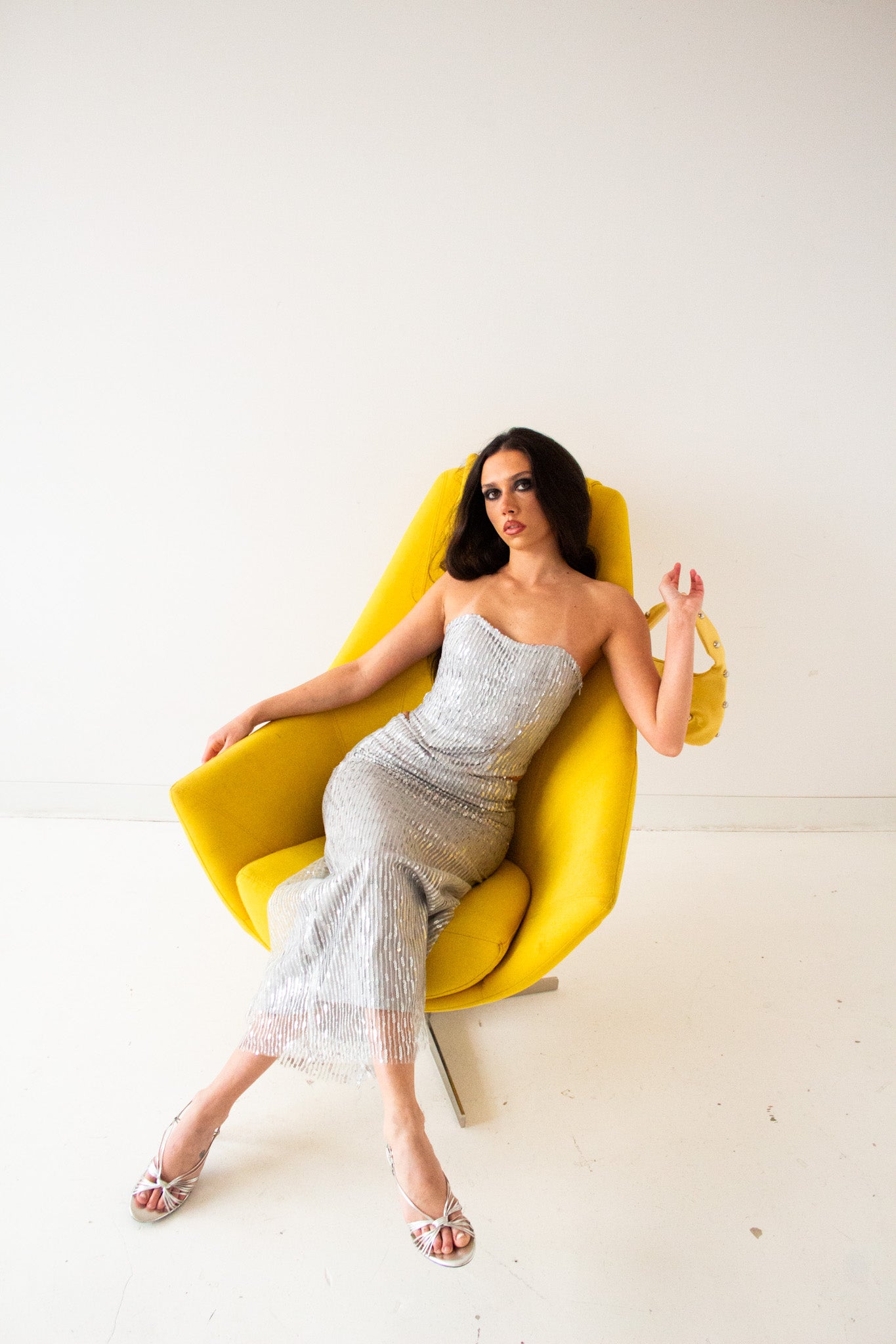 Model in a strapless silver sequin set, reclining back in a yellow sculptural chair, holding a yellow studded handbag. Editorial, chic, and confident studio vibe.