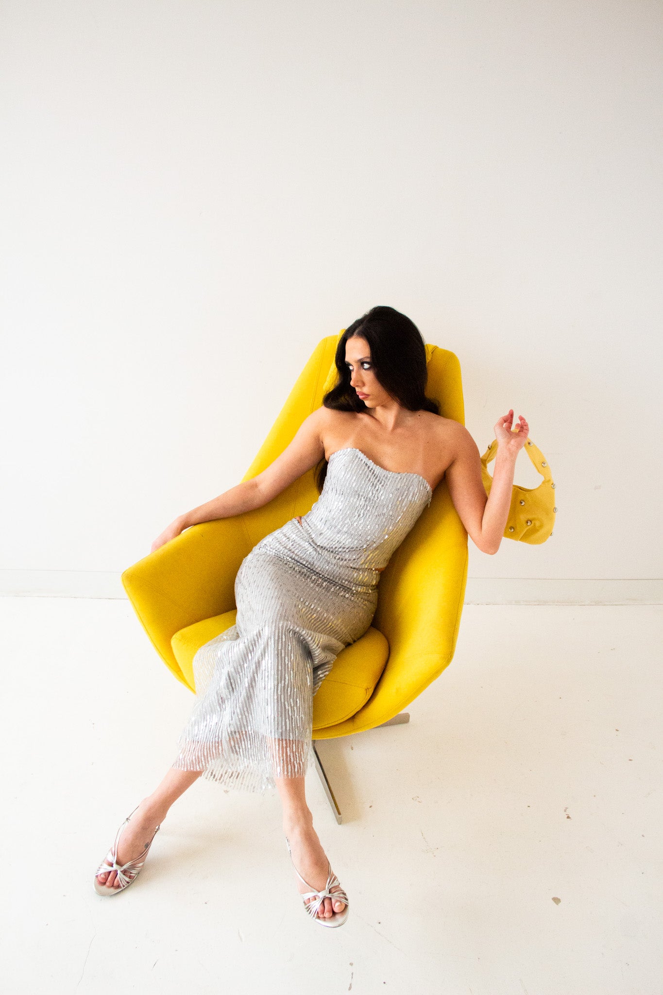 Model in a silver sequin matching set, flipping her hair with attitude while posing in a yellow designer chair, silver heels completing the look. Shot in a clean, minimalist studio.
